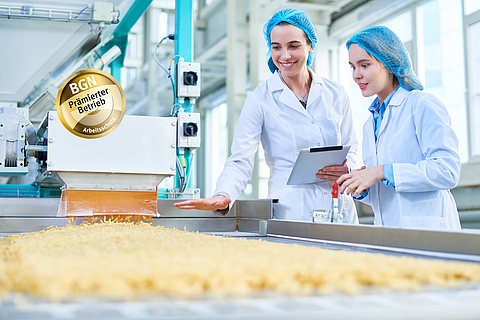 Zwei Arbeiterinnen mit Schutzkleidung stehen neben einem Laufband mit Nudeln in einer Produktionshalle.