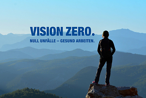 Eine Person steht auf einem Berggipfel und schaut in die Ferne. Daneben steht "Vision Zero. Null Unfälle - gesund arbeiten."