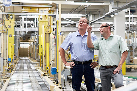 Zwei Männer stehen in einer Fabrikhalle, ein Mann zeigt auf etwas.