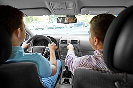 Blick von hinten im Auto auf Fahrer und Beifahrer, der Beifahrer erklärt dem Fahrer etwas.
