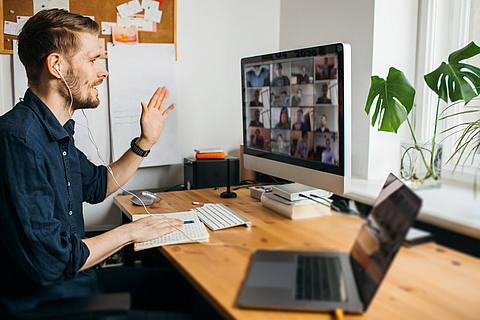 Ein Mann sitzt am Schreibtisch vor einem PC, auf dem Bildschirm läuft ein Videochat. Der Mann winkt in die Kamera.
